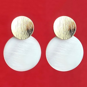 Puede incluir: Un par de pendientes de oro y blanco. Los pendientes están hechos de dos círculos, un círculo dorado más pequeño encima de un círculo blanco más grande.