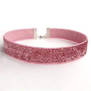 Könnte beinhalten: Ein rosafarbener Samt-Choker mit einem silbernen Verschluss. Der Choker hat eine glitzernde, strukturierte Oberfläche.