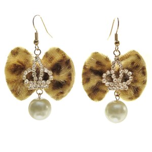 Peut inclure: Une paire de boucles d'oreilles dorées avec un nœud en fausse fourrure et un charme de couronne en strass. Chaque boucle d'oreille est rehaussée d'une perle blanche.