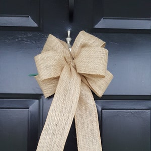 May include: A large, rustic burlap bow with a natural, beige color. The bow is tied with a loop and hangs from a door.