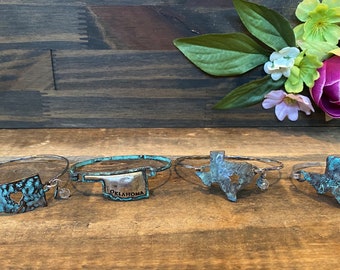 May include: Five silver bracelets with state-shaped charms. The charms are in the shape of Oklahoma, Texas, and a heart-shaped cutout of Oklahoma. The charms are a turquoise blue color with a distressed finish.