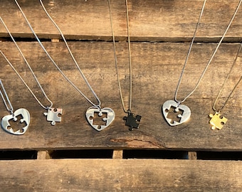 May include: Five silver necklaces with puzzle piece pendants. Two pendants are heart-shaped with a puzzle piece cutout, one is a solid puzzle piece, and two are solid puzzle pieces in black and gold.