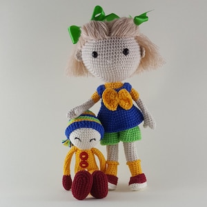 Könnte beinhalten: Zwei gehäkelte Puppen, eine steht und eine sitzt. Die stehende Puppe hat blonde Haare, ein blaues und gelbes Kleid, grüne Shorts und rote Schuhe. Die sitzende Puppe hat einen gelben Körper, eine blaue und grüne Mütze und rote Schuhe.