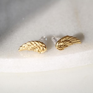 Könnte beinhalten: Ein Paar goldfarbene Ohrstecker in Flügelform. Die Ohrringe bestehen aus einem glänzenden, metallischen Material und sind auf einer weißen Marmoroberfläche platziert. Die Ohrringe haben ein detailliertes Federdesign.