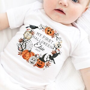 May include: White baby onesie with a Halloween-themed wreath design. The wreath includes pumpkins, a crow, a spiderweb, and the text "MY FIRST HALLOWEEN Elsie 2023". The baby is wearing an orange bow.