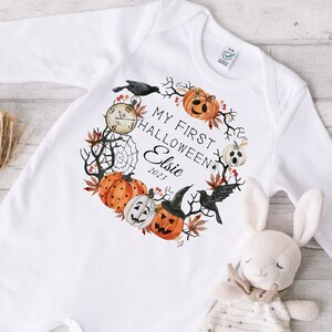 May include: White baby onesie with long sleeves, featuring a Halloween-themed wreath design. The wreath includes pumpkins, a clock, a spiderweb, and the text "My First Halloween Elsie 2023". A stuffed bunny and a bundle of twine are also in the image.
