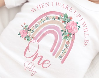 May include: White baby onesie with a pink and peach rainbow with floral accents. The text "When I wake up I will be one Tilly" is printed on the onesie.