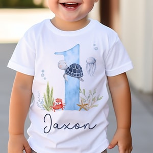 K&ouml;nnte beinhalten: Ein wei&szlig;es T-Shirt mit einer blauen Zahl Eins darauf. Die Eins ist mit einer Meeresschildkr&ouml;te, einer Qualle, einem Krebs und einem Seestern verziert. Der Text "Jaxon" ist unter der Eins gedruckt.