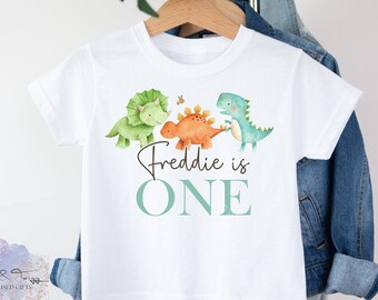 May include: White t-shirt with a green, orange, and blue dinosaur design. The text "Freddie is ONE" is printed in brown and teal.