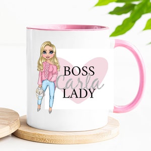 May include: White ceramic mug with a pink rim and handle. The mug features a cartoon illustration of a woman with blonde hair and a pink shirt. The text "BOSS Carla LADY" is printed on the mug in a pink heart.