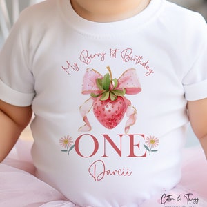 May include: White short-sleeved t-shirt with a strawberry design and the text "My Berry 1st Birthday". The design includes a pink bow, the word "ONE", and the name "Darcii". The shirt is for a baby.