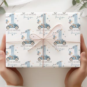 May include: White wrapping paper featuring a repeating pattern of a blue race car with the number 1, balloons, and stars. The text "HAPPY 1ST BIRTHDAY George" is printed on the paper. The paper is tied with twine.