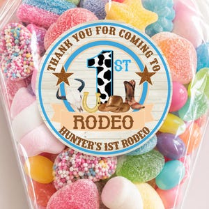 Peut inclure: Un sachet en plastique transparent rempli de bonbons gélifiés colorés, avec un autocollant circulaire sur fond imitation bois. L'autocollant porte le texte "THANK YOU FOR COMING TO 1ST RODEO" et "HUNTER'S 1ST RODEO" avec un motif de vache, un crâne, un fer à cheval et des bottes.