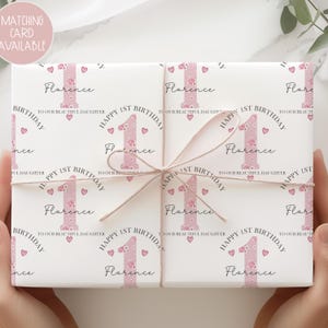 May include: A wrapped gift with a pink floral "1" design, the name "Florence", and the text "Happy 1st Birthday". The gift is tied with twine and features a matching card available. The wrapping paper has a white background with pink hearts.