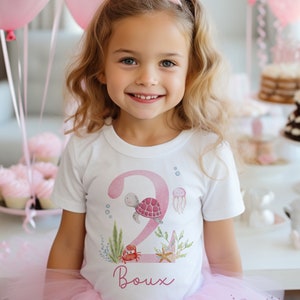 K&ouml;nnte beinhalten: Ein wei&szlig;es T-Shirt mit einer pinkfarbenen Zahl Zwei, einer Schildkr&ouml;te, einem Krebs, einem Seestern und Seegras. Der Name "Boux" steht unter der Zahl. Das Shirt wird von einem Kind getragen, das einen rosa T&uuml;llrock und eine rosa Partym&uuml;tze tr&auml;gt.