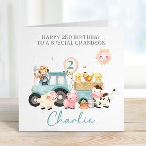 May include: A birthday card with a farm theme. The card features a light blue tractor, a sheep, a pig, a cow, and several chicks. The text reads "Happy 2nd Birthday to a Special Grandson" and "Charlie". A smiling sun is also included.