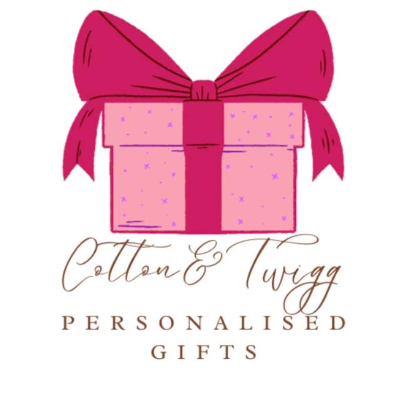 May include: A pink gift box with a magenta bow and ribbon. The box is decorated with small purple crosses. The text "Cotton & Twigg" is written in a cursive font above the words "Personalised Gifts".