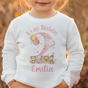 May include: White long-sleeved shirt with a pink watercolour number two, a sun, a rainbow, a cloud, and a line of animals. The text "It's my Birthday" is above the number and the name "Emilia" is below the number.