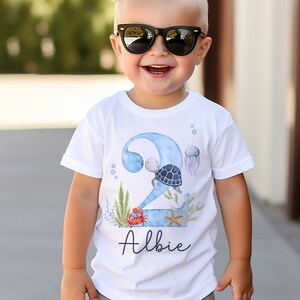 K&ouml;nnte beinhalten: Wei&szlig;es T-Shirt mit einer blauen Zahl Zwei, einer Schildkr&ouml;te, einem Krebs, einem Seestern und Seegras. Der Name "Albie" steht unter der Zahl.