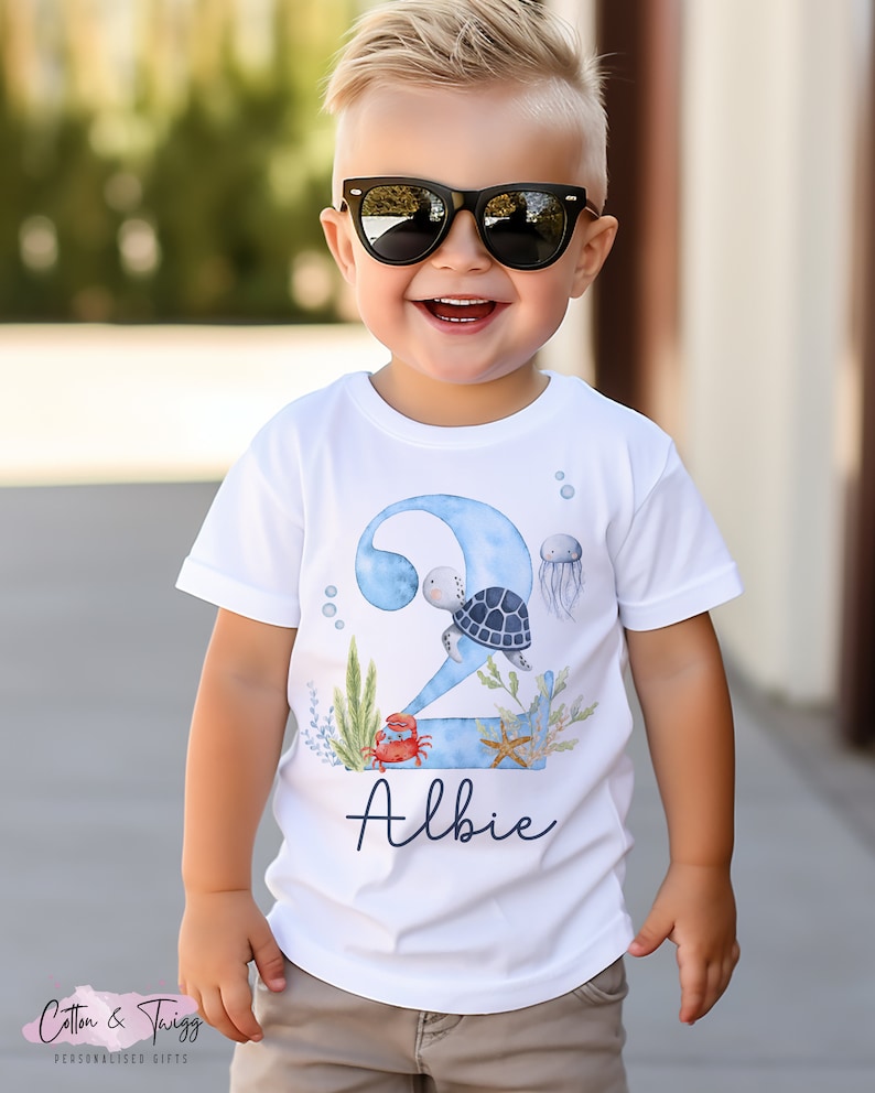 K&ouml;nnte beinhalten: Wei&szlig;es T-Shirt mit einer blauen Zahl Zwei, einer Schildkr&ouml;te, einem Krebs, einem Seestern und Seegras. Der Name "Albie" steht unter der Zahl.
