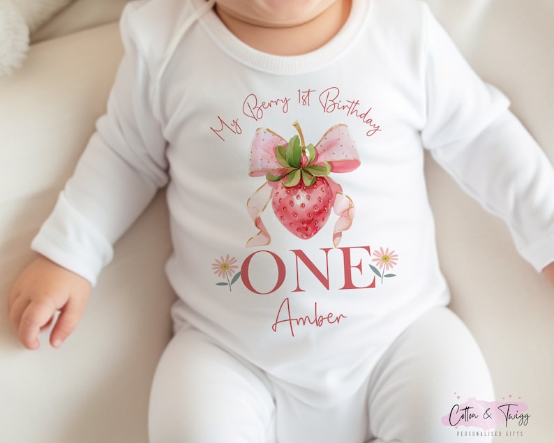 May include: White long-sleeved baby bodysuit with a strawberry design. The design includes a pink strawberry with a bow, the text "My Berry 1st Birthday", the word "ONE", and the name "Amber".