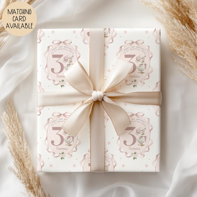 May include: A wrapped gift with a light pink ribbon and bow. The wrapping paper features a repeating pattern of a pink oval with a number 3, a cartoon duck, and the text "Happy 3rd Birthday to my beautiful niece Florence". A matching card is available.