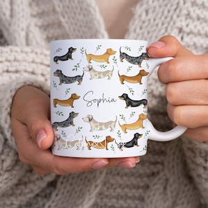 Puede incluir: Taza de cer&aacute;mica blanca con un patr&oacute;n de perros salchicha en varios colores y poses. La taza tiene un asa negra y el nombre "Sophia" est&aacute; escrito en letras negras en la parte delantera.