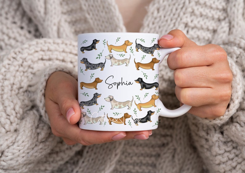 Puede incluir: Taza de cer&aacute;mica blanca con un patr&oacute;n de perros salchicha en varios colores y poses. La taza tiene un asa negra y el nombre "Sophia" est&aacute; escrito en letras negras en la parte delantera.