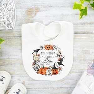 May include: White baby bib with a Halloween-themed design. The bib features a wreath of pumpkins, crows, and a spiderweb around the text "MY FIRST HALLOWEEN Elsie 2023". The design includes a clock and a jack-o'-lantern wearing a witch hat.