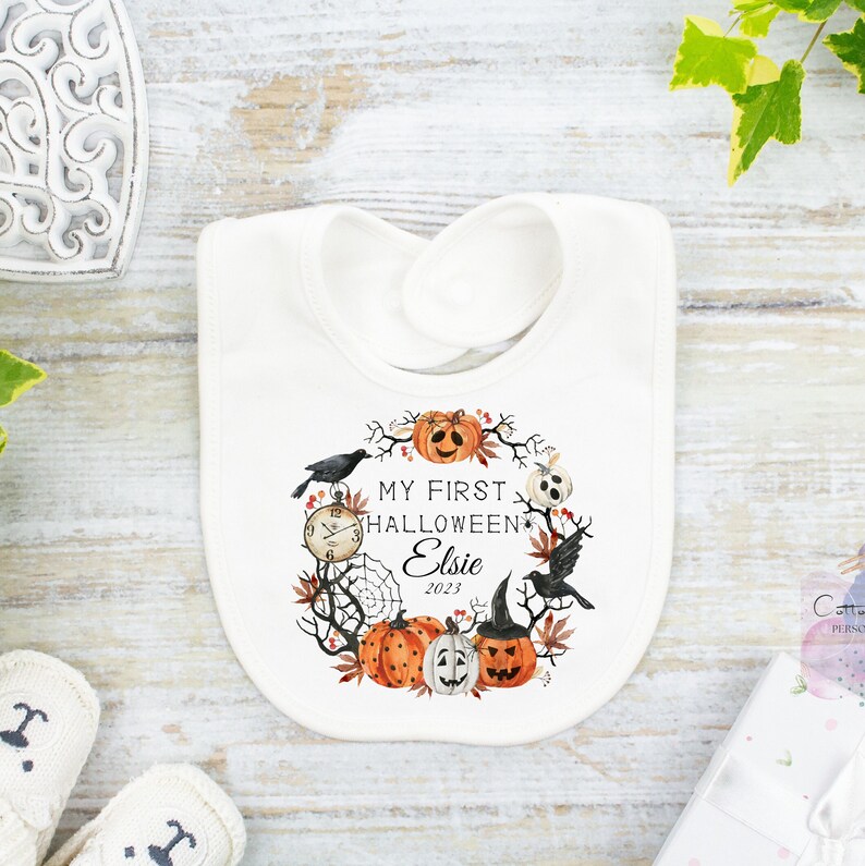 May include: White baby bib with a Halloween-themed design. The bib features a wreath of pumpkins, crows, and a spiderweb around the text "MY FIRST HALLOWEEN Elsie 2023". The design includes a clock and a jack-o'-lantern wearing a witch hat.