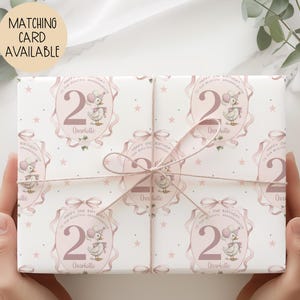 May include: Gift-wrapped present featuring a repeating pattern of a duck wearing a party hat, the number 2, and the name "Charlotte" on a white background. The wrapping paper is tied with a pink ribbon. Text on the wrapping paper reads "Happy 2nd Birthday to our beautiful granddaughter."
