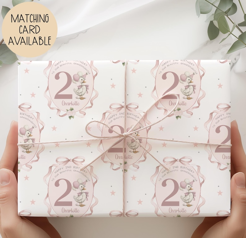 May include: Gift-wrapped present featuring a repeating pattern of a duck wearing a party hat, the number 2, and the name "Charlotte" on a white background. The wrapping paper is tied with a pink ribbon. Text on the wrapping paper reads "Happy 2nd Birthday to our beautiful granddaughter."