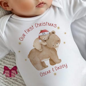 Puede incluir: Body de bebé blanco con una ilustración de dos osos. El oso más grande lleva un oso más pequeño con un gorro de Papá Noel con "2025". El texto dice "Our First Christmas" y "Oscar & Daddy" en rojo, con estrellas.
