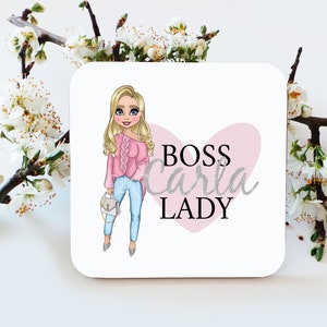 May include: A white square coaster with a cartoon illustration of a woman with blonde hair wearing a pink sweater and blue jeans. The text "BOSS Carla LADY" is written in silver glitter font on a pink heart.