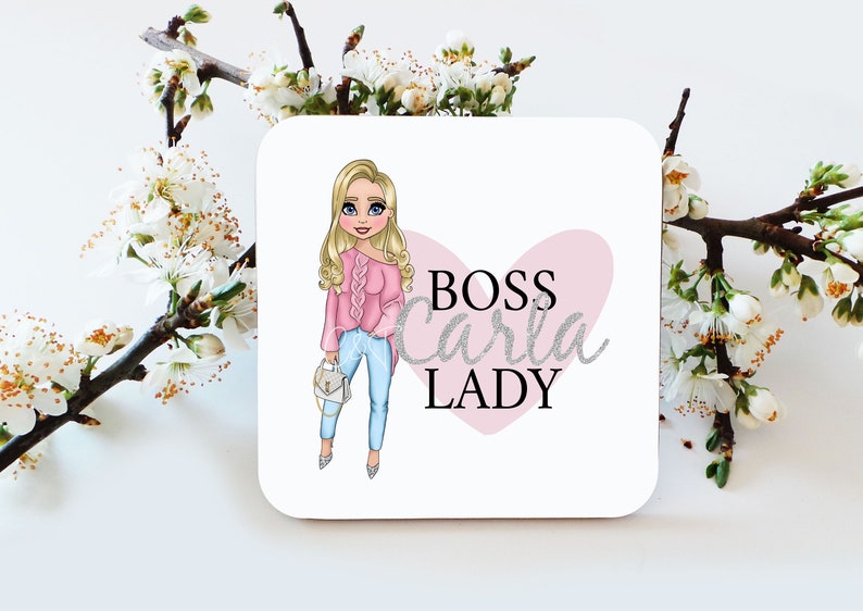May include: A white square coaster with a cartoon illustration of a woman with blonde hair wearing a pink sweater and blue jeans. The text "BOSS Carla LADY" is written in silver glitter font on a pink heart.