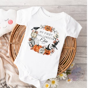 May include: White baby onesie with a Halloween-themed wreath design. The wreath includes pumpkins, a crow, a spiderweb, and the text "My First Halloween Elsie 2023". The onesie is placed on a wicker basket.