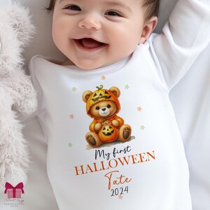 Puede incluir: Body de bebé blanco con un diseño de Halloween. El diseño presenta un osito de peluche disfrazado de calabaza, sosteniendo una calabaza. El texto en el body dice "My first Halloween" y "Tate 2024".