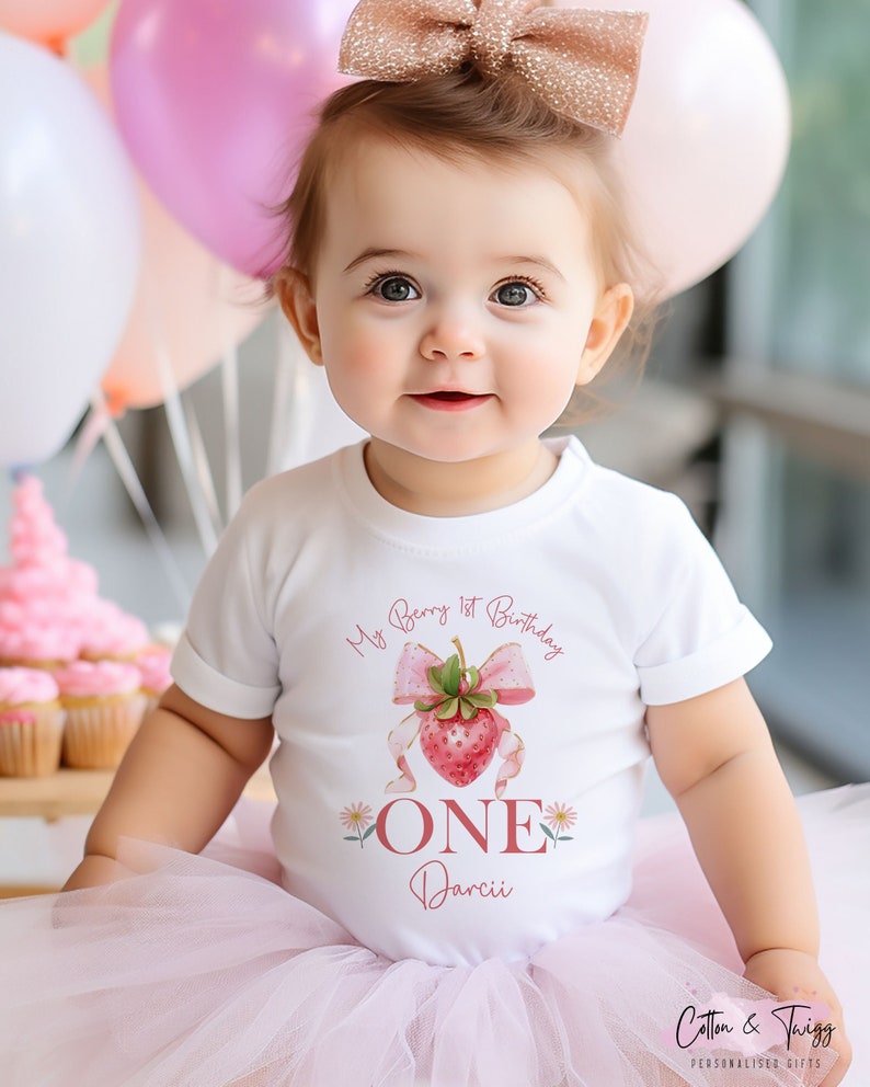 May include: A baby wearing a white t-shirt with a strawberry graphic and the text "My Berry 1st Birthday ONE Darcii". The baby is wearing a pink tutu and a rose gold bow. Pink and white balloons are in the background.