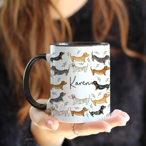 Puede incluir: Taza de cer&aacute;mica blanca con asa y borde negros. La taza presenta un patr&oacute;n de perros salchicha de varios colores y poses, con hojas verdes. El nombre "Karen" est&aacute; escrito en cursiva negra en la taza.