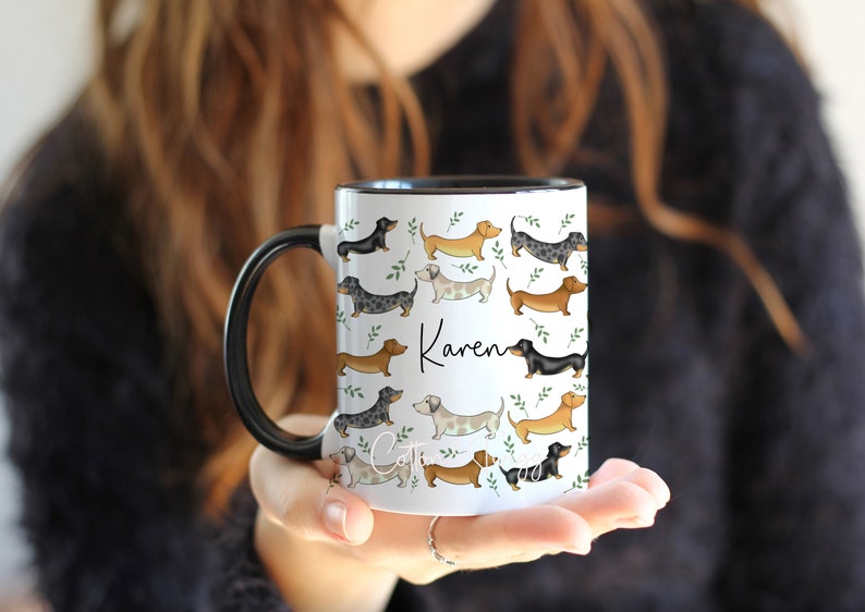 Puede incluir: Taza de cer&aacute;mica blanca con asa y borde negros. La taza presenta un patr&oacute;n de perros salchicha de varios colores y poses, con hojas verdes. El nombre "Karen" est&aacute; escrito en cursiva negra en la taza.
