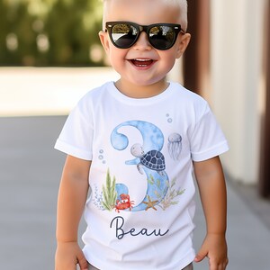 K&ouml;nnte beinhalten: Wei&szlig;es T-Shirt mit einer blauen Aquarell-Nummer 3, einer Schildkr&ouml;te, einer Qualle, einem Krebs und einem Seestern. Das Shirt tr&auml;gt auch den Namen "Beau" aufgedruckt.
