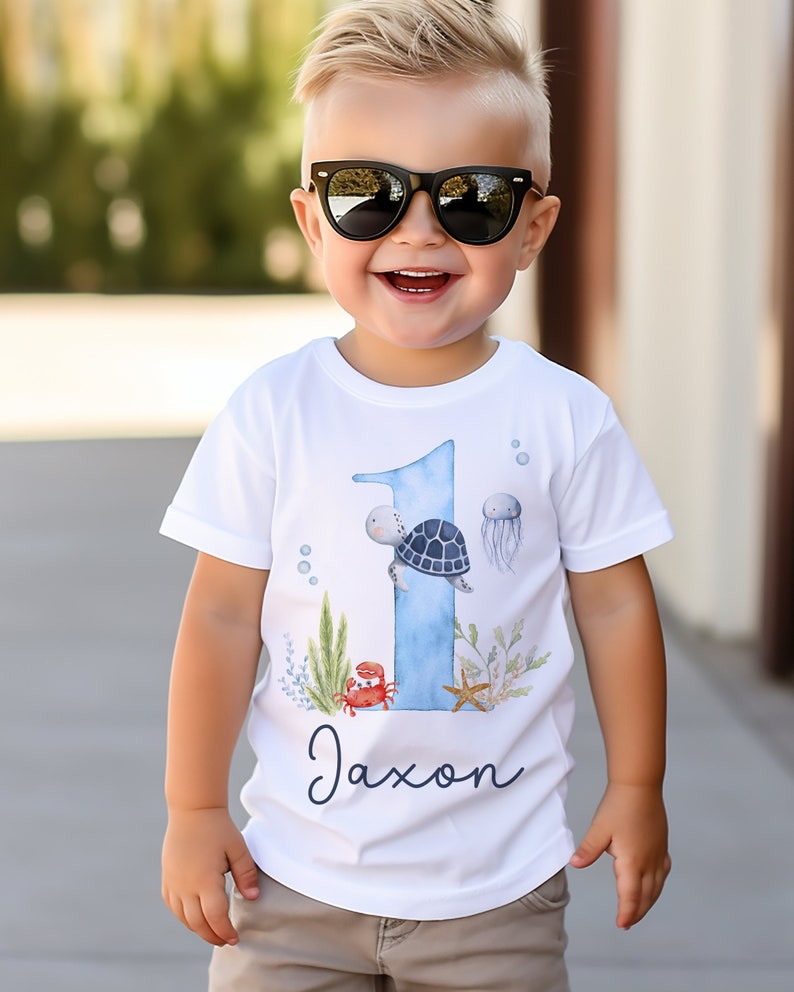 K&ouml;nnte beinhalten: Ein wei&szlig;es T-Shirt mit einer blauen Zahl Eins darauf. Die Eins ist mit einer Meeresschildkr&ouml;te, einer Qualle, einem Krebs und einem Seestern verziert. Der Text "Jaxon" ist unter der Eins gedruckt.