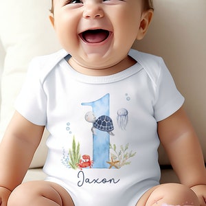 K&ouml;nnte beinhalten: Wei&szlig;er Baby-Strampler mit einer blauen Zahl eins, einer Schildkr&ouml;te, einem Krebs, einem Seestern und einer Qualle. Der Strampler hat den Namen Jaxon darauf gedruckt.