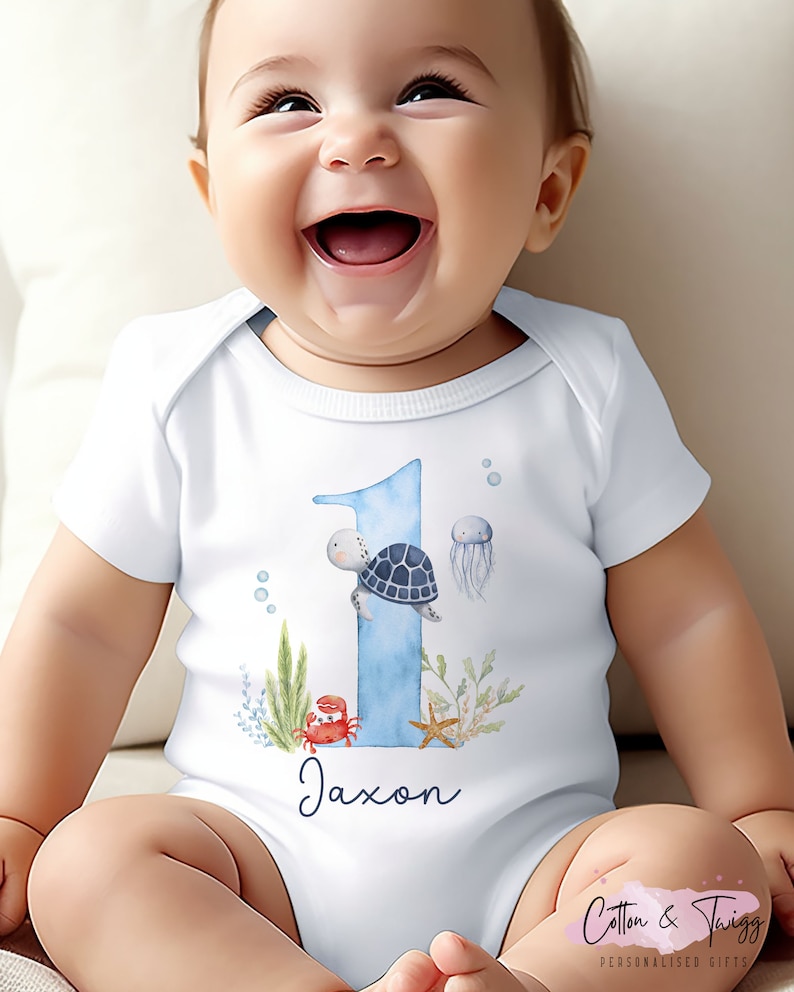 K&ouml;nnte beinhalten: Wei&szlig;er Baby-Strampler mit einer blauen Zahl eins, einer Schildkr&ouml;te, einem Krebs, einem Seestern und einer Qualle. Der Strampler hat den Namen Jaxon darauf gedruckt.