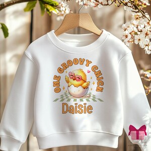 Puede incluir: Sudadera blanca con un gráfico de un pollito amarillo con gafas de sol y el texto "One Groovy Chick Daisie".
