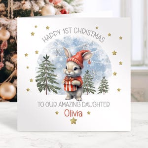 May include: A Christmas card with a watercolor illustration of a bunny wearing a red hat and sweater, holding a gift. The card reads "Happy 1st Christmas To Our Amazing Daughter Olivia" with gold star accents.