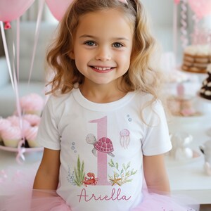 K&ouml;nnte beinhalten: Ein wei&szlig;es T-Shirt mit einer pinkfarbenen Zahl eins, einer Schildkr&ouml;te, einer Qualle und einem Krebs. Das Shirt tr&auml;gt auch den Namen "Ariella" aufgedruckt.