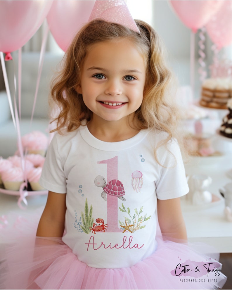 K&ouml;nnte beinhalten: Ein wei&szlig;es T-Shirt mit einer pinkfarbenen Zahl eins, einer Schildkr&ouml;te, einer Qualle und einem Krebs. Das Shirt tr&auml;gt auch den Namen "Ariella" aufgedruckt.