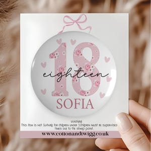 Puede incluir: Un botón redondo rosa y blanco con el número "18" y la palabra "eighteen" en cursiva. El número está decorado con motivos florales. El nombre "SOFIA" está debajo. Pequeños corazones rosas están dispersos alrededor del botón.
