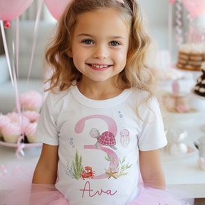 K&ouml;nnte beinhalten: Ein wei&szlig;es T-Shirt mit einer pinkfarbenen Zahl 3, einer Schildkr&ouml;te, einem Krebs, einem Seestern und Algen. Der Name Ava ist in Pink unter der Zahl geschrieben.